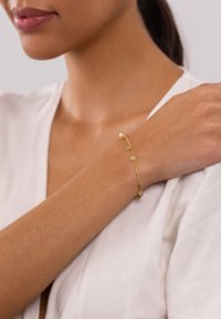 Goldarmband an einem hellhäutigen Arm. Das Design zeigt kleine zirkuläre Akzente, die durch eine filigrane Kette verbunden sind. Minimalistischer und eleganter Stil.