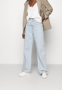 Person som bär en beige kavaj över en vit spetstopp, ljusblå vida jeans och vita sneakers, som står framför en enfärgad bakgrund.
