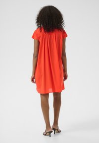 Kurzes oranges Baumwollkleid mit gerafftem Rückendetail, lockerer Passform und knielanger Länge. Tragen mit schwarzen Riemchensandalen mit hohen Absätzen.