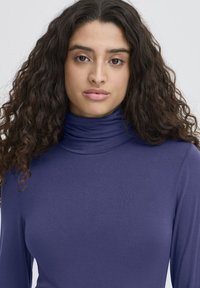 Giovane donna con lunghi capelli ricci e scuri, che indossa un maglione a collo alto viola, mentre guarda direttamente nella fotocamera con un'espressione neutra.