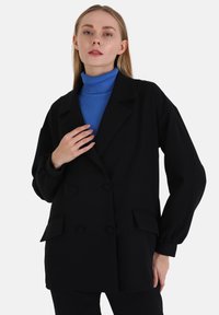 Zwarte dubbelknopen blazer met gepelde revers, twee voorzakken en lange mouwen. Draag over een blauwe ribbelige coltrui.