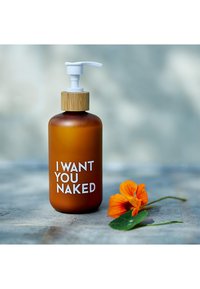 Amber glazen fles met een bamboe pompdeksel; bevat witte tekst met de zin "IK WIL JE NAakt." Oranje bloem en groene bladeren op de voorgrond.