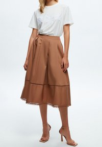 T-shirt blanc avec des motifs floraux brodés, associé à une jupe marron à silhouette évasée et ourlet festonné. Sandales à talons carrés beige.