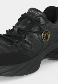 Gros plan d'une chaussure de sport noire avec une semelle épaisse, des lacets noirs, une tige texturée et un emblème métallique rond présentant deux oiseaux dorés.