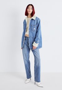 Denimjack met een crèmekleurige nepshearlingkraag, blauwe vervaagde kleur, knopen aan de voorkant en twee borstzakken; gecombineerd met een spijkerbroek en witte hakken.