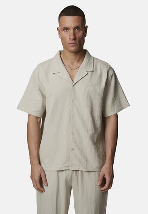 Uomo con capelli corti che indossa una camicia beige a maniche corte con bottoni e pantaloni abbinati, in piedi davanti a uno sfondo semplice.