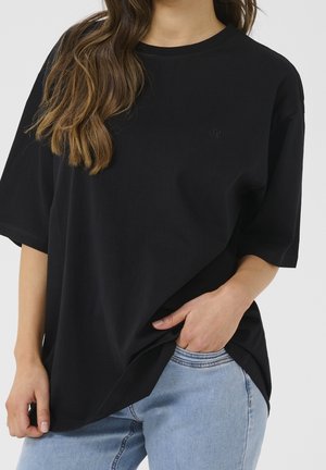 Schwarzes, übergroßes T-Shirt aus Baumwolle mit kurzen Ärmeln, Rundhalsausschnitt und einem dezenten Logodetail auf der Brust. Kombiniert mit hellblauen Jeans.