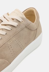Beige semsket sneakers med avrundet tå, kremfargede snørebånd og subtil perforert detaljering. Har en hvit gummisåle og preget logo.