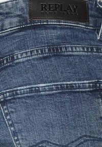 Sininen denim-farkut, joissa on sileä rakenne, musta nahkapläntti, jossa lukee "REPLAY BLUE JEANS", sekä näkyvät ompelualueet.