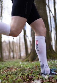 Kompressionsstrümpfe in Weiß mit schwarzen Akzenten, versehen mit einem Logo, einem strukturierten Grip-Band am oberen Ende und einem Verlaufstreifenmuster.