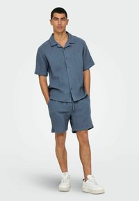 Korte mouw button-up shirt en short set in gestructureerde blauwe stof, met een kraag, vooraan knopen en een elastische tailleband. Witte sneakers.
