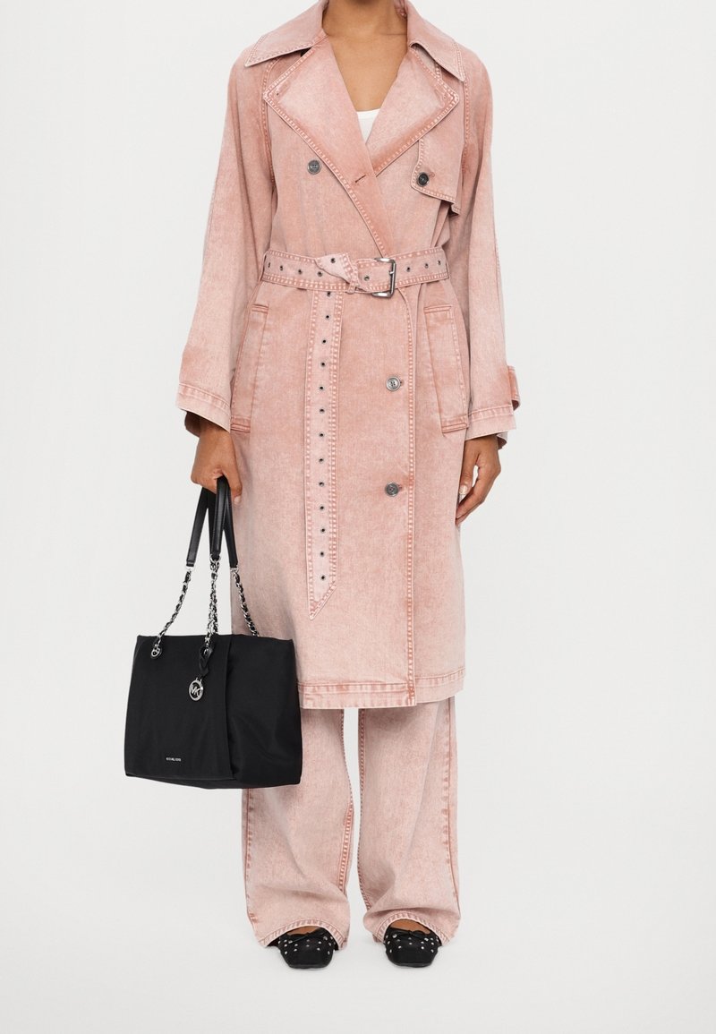 Femme portant un trench-coat rose délavé ceinturé et un pantalon à jambes larges assorti, tenant un sac à main noir avec des anses en chaîne.