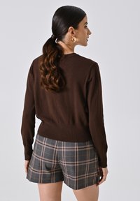 Maglione lavorato a maglia marrone con scollatura rotonda, abbinato a pantaloni corti a quadri grigi con accenti arancioni e bianchi. Il maglione ha un bordo a costine.