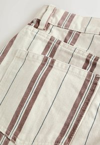 Gros plan d'un tissu beige avec des rayures verticales marron et fines noires, montrant une poche arrière cousue et un détail de passant de ceinture.