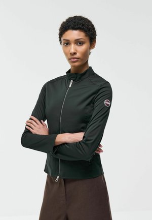 Osoba z krótkimi kręconymi włosami, ubrana w czarną kurtkę na zamek z naszywką z logo na górnej części rękawa, stojąca z założonymi skrzyżowanymi ramionami.