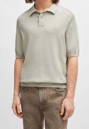 Person trägt ein hellgraues, kurzärmeliges Strick-Poloshirt mit zwei Knöpfen und "BOSS"-Logo, kombiniert mit braunen Jeans aus Denim.