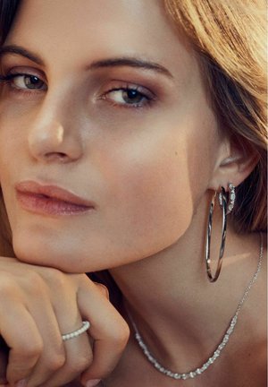 Sølv hoop øreringe, en ring med krystaller og en delikat halskæde med klare sten. Huden ser glat ud med naturlige makeup nuancer.