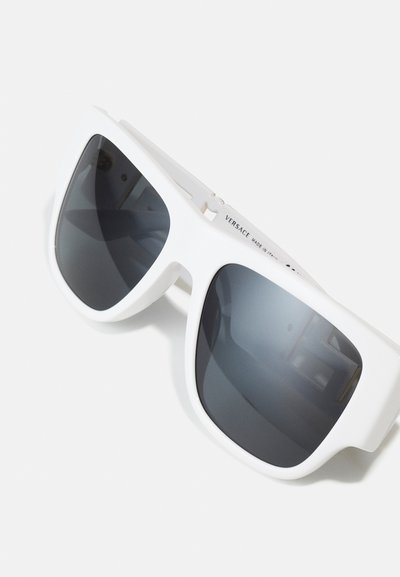 Lunettes de soleil blanches surdimensionnées avec des verres sombres, monture en plastique épaisse et une étiquette de marque bien visible sur le côté. Texture lisse et design angulaire.