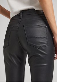 Detalle en primer plano de una persona que lleva jeans negros con un acabado mate y bolsillos traseros, con una parte superior blanca, mostrando detalles de costura y textura.