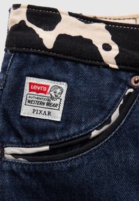 Levi's farkut esittelevät sinistä kangasta, jossa on musta ja kerma-valkoinen lehmäkuvioinen vyötärö. Valkoisessa tarrassa lukee "Aito lännen muoti" ja "Pixar."