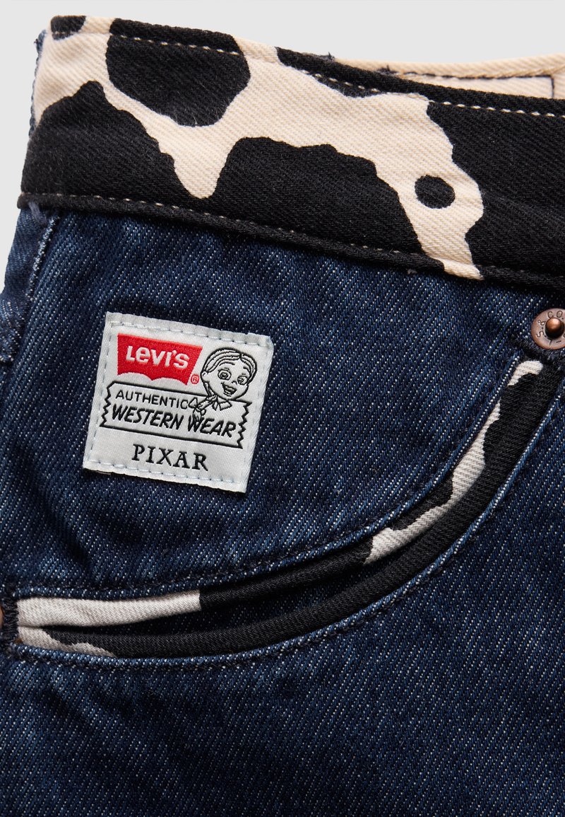 Levi's denim jeans har en blå stof med en sort og creme cow print talje. Et hvidt mærke viser "Authentic Western Wear" og "Pixar."