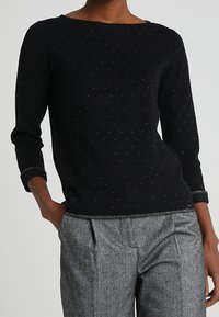 Maglione nero con scollo a barca e maniche a tre quarti, caratterizzato da un motivo testurizzato di piccole pois argentate. Abbinato a pantaloni grigi.