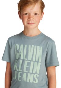Camiseta de algodón de color azul claro con gran texto verde "CALVIN KLEIN JEANS." Manga corta, escote redondo y corte relajado.