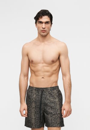 Hea sportliku kehaehitusega mees tume juustega, kes kannab halli madu mustriga ujumispükse, seisab ühtlase heleda taustaga vastu ja vaatab ette.