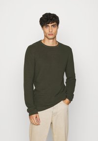 Mørkegrønn ribbestrikket genser med rund hals og lange ermer, med et teksturert mønster. Iført lysebeige bukser.