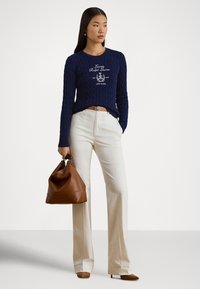 Mornarsko modri pulover z vzorcem kablovskega pletenja in logotipom spredaj, kombiniran s kremasto širokimi hlačami in rjavo usnjeno torbico; modelka drži torbico z eno roko.