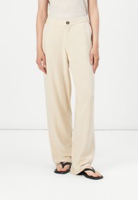 Pantaloni beige a gamba larga in un tessuto leggero con una trama liscia, dotati di chiusura a bottone e tasche laterali. Sandali piatti completano il look.