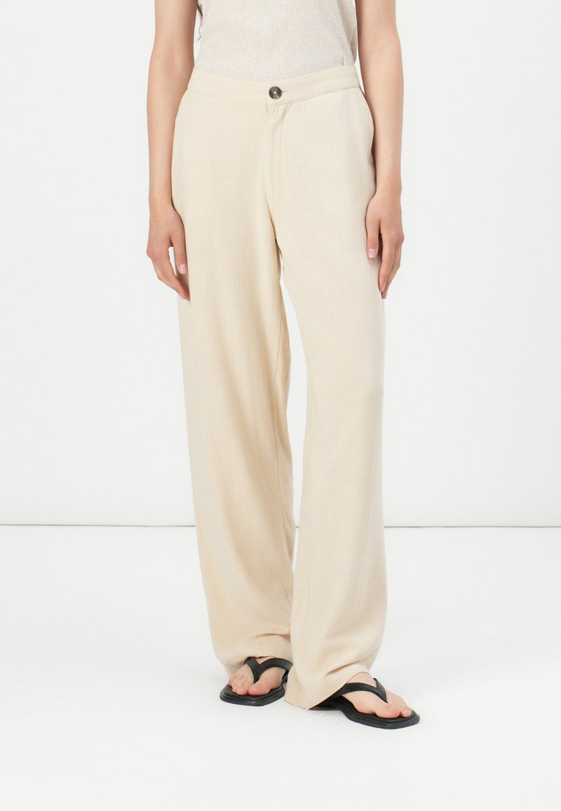 Pantaloni beige a gamba larga in un tessuto leggero con una trama liscia, dotati di chiusura a bottone e tasche laterali. Sandali piatti completano il look.