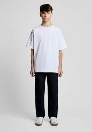 Tienerjongen staand, draagt een effen wit oversized T-shirt, zwarte broek en witte sneakers tegen een grijze achtergrond.