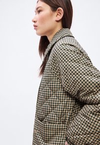 Prošívaná bunda s motivem psa zubatého v zelené a béžové barvě, s límcem a viditelnými bočními kapsami, zobrazená na profilu modelky.