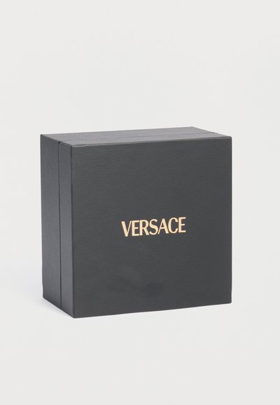 Μαύρο ορθογώνιο κουτί με χρυσό λογότυπο «VERSACE» στο κέντρο της μιας πλευράς, σε απλό ανοιχτό γκρι φόντο.