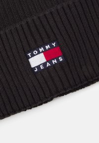Crni rebrasti zimski šešir s pravokutnom tkaninom na kojoj je logo "TOMMY JEANS" u bijelim, plavim i crvenim sekcijama. Mekana pletena tekstura.