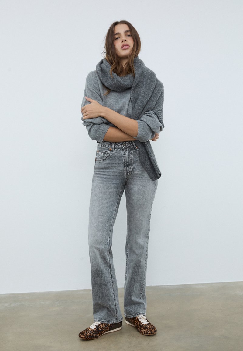 Grå sweater lagd over en grå top, parret med lysegrå flared jeans og sneakers med leopardprint, mod en helt hvid baggrund.