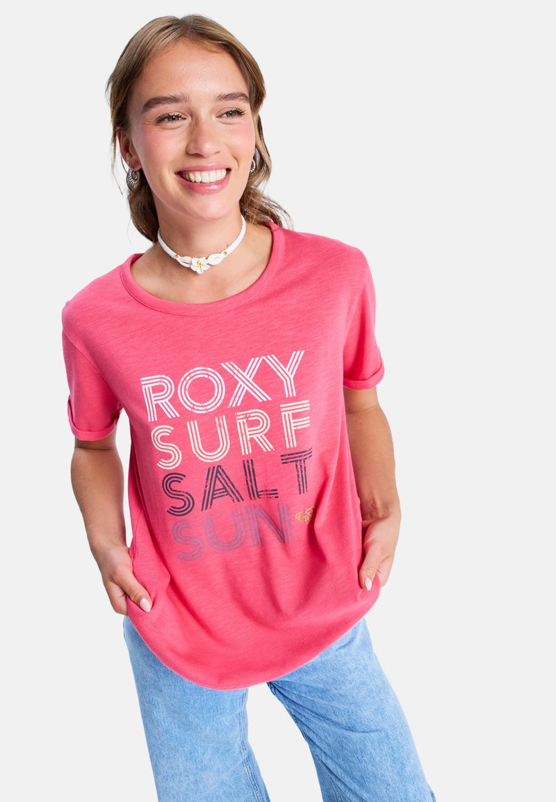 Smilende kvinde iført en pink "ROXY SURF SALT SUN" T-shirt, lyseblå jeans, muslinghalskæde og sølv hoop øreringe, hænderne i lommerne.