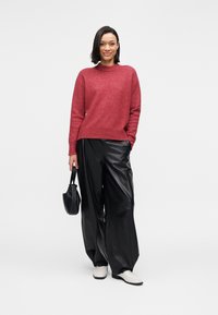 Pull rouge en tricot avec un col rond, associé à un pantalon large en cuir noir. Des chaussures blanches et un petit sac noir complètent la tenue.