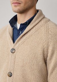 Man in een beige gebreide cardigan met twee knopen over een donkerblauw overhemd met knopen en een wit onderhemd.