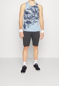 Camiseta sin mangas azul para hombre con patrón de montaña, pantalones cortos grises, pulseras blancas, zapatos negros con acentos naranjas, diseño atlético.