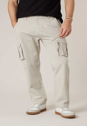 Homme portant un pantalon cargo beige avec poches zippées et à rabat, des baskets blanches, un t-shirt noir et des bracelets de perles, les mains dans les poches.