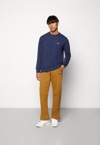 Navy sweatshirt with small multi-colored logo, paired with mustard sweatpants and white sneakers. Smooth fabric with a relaxed fit design.