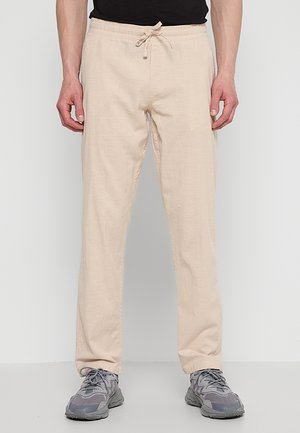 Mężczyzna w beżowych spodniach ze sznurkiem i szarych trampkach stoi na jednolitym jasnym tle.