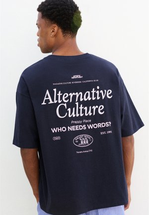 Homme portant un T-shirt bleu marine avec un grand texte blanc et rose indiquant "Alternative Culture" et "Who needs words ?" dans le dos.