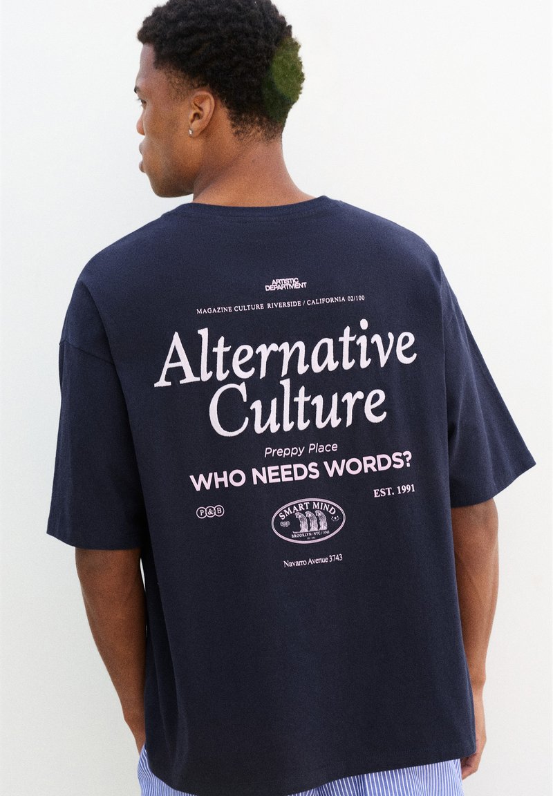 Homme portant un T-shirt bleu marine avec un grand texte blanc et rose indiquant "Alternative Culture" et "Who needs words ?" dans le dos.