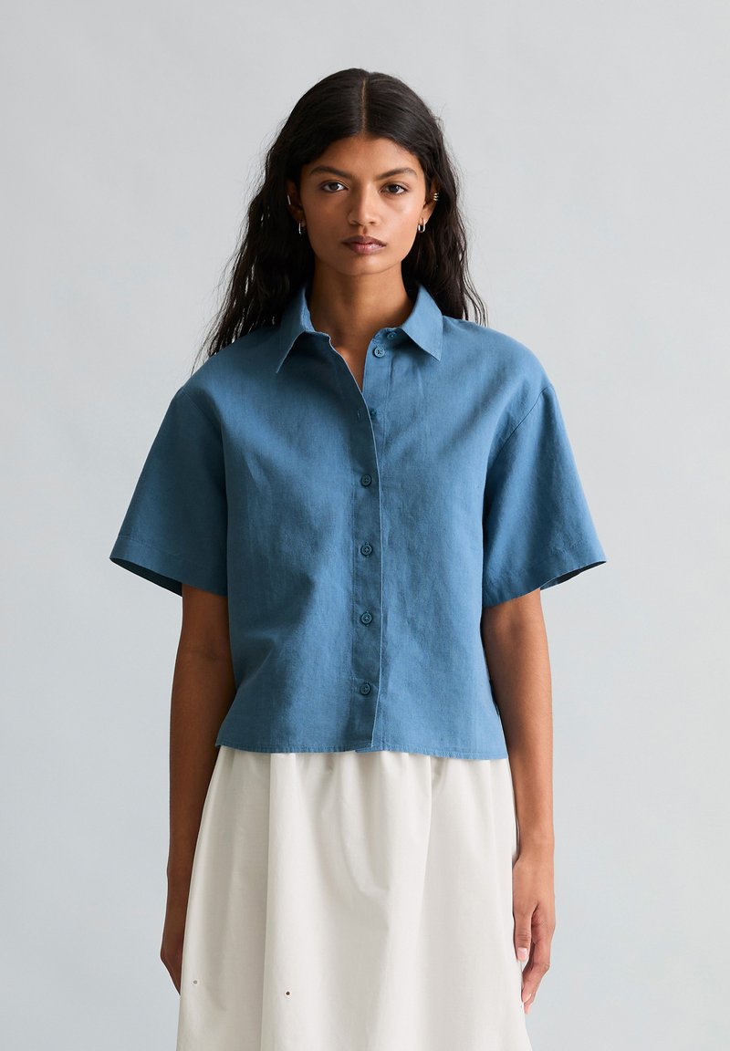 Femme aux longs cheveux foncés portant une chemise bleue ample à manches courtes boutonnée et une jupe blanche, debout devant un fond gris clair uni.