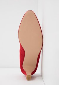 Chaussure à talon haut en daim rouge avec un bout pointu, une semelle beige lisse et des détails texturés le long des bords.