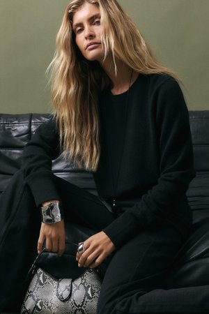 Model oblečený v čiernom svetri a nohaviciach, drží čiernu koženú tašku s hadím vzorom. Na zápästiach a prstoch má strieborné šperky.