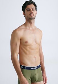 Heren fitted boxershorts in olijfgroen met een zwarte tailleband waarop witte "LACOSTE" branding staat. Soepele stof, minimalistisch ontwerp.